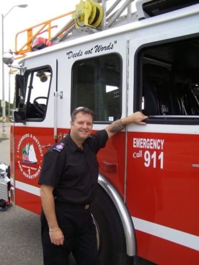 A Lunenburg and District firefighter in duty uniform beside the department's ladder truck, marked Deeds not Words